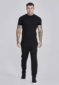 Juodas trumpomis rankovėmis sportinis marškinėlis su tekstūruotu kaklu, derinamas su juodomis siaurėjančiomis trikotažinėmis kelnėmis, ant kurių yra mažas logotipas ir šoniniai kišeniai.