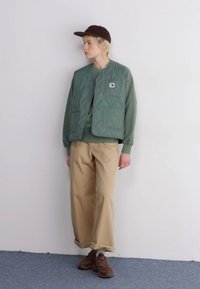 Gilet trapuntato verde sopra un maglione verde coordinato, pantaloni beige a gamba larga, cappello marrone e sneakers marroni. Texture liscia con cuciture visibili.