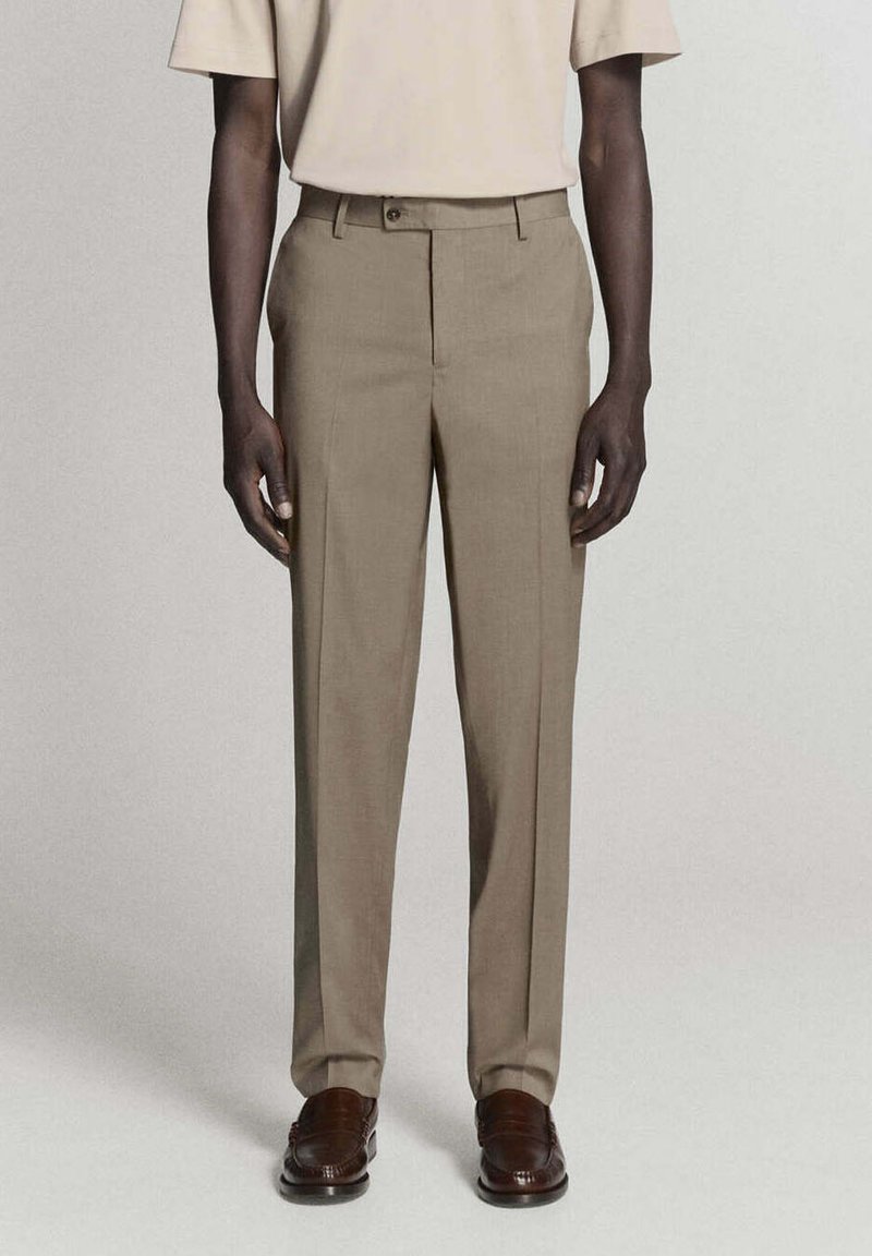 Personne debout, portant une chemise beige à manches courtes, un pantalon de costume beige et des chaussures habillées marron sur un fond clair et uni.