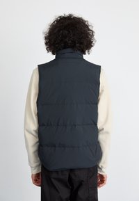 Colete puffer preto com gola alta e padrão acolchoado visível. Usado sobre uma camisa de manga longa bege clara, exibindo um corte descontraído.