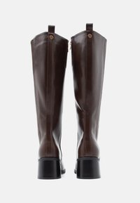 Καφετινά δερμάτινα μποτάκια που φτάνουν μέχρι το γόνατο με τετράγωνη φτέρνα, διαθέτουν κλείσιμο με φερμουάρ και δύο διακοσμητικά κουμπιά στην κορυφή. Λείος υφασμάτινος σχεδιασμός.