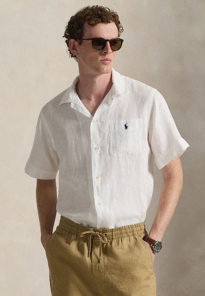 Chemise en lin blanche à manches courtes avec une poche poitrine, design boutonné, associée à un short marron clair à cordon de serrage et des lunettes de soleil foncées.