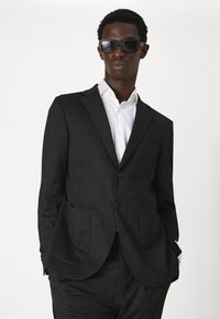 Veste de costume à carreaux sombres avec deux boutons, revers crantés et poches plaquées. Une chemise blanche en dessous et des lunettes de soleil noires complètent le look.