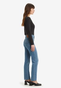 Jeans de ganga azul com cintura alta, corte reto, superfície texturizada e um remendo traseiro castanho em contraste; combinados com um top preto de mangas compridas ajustado.