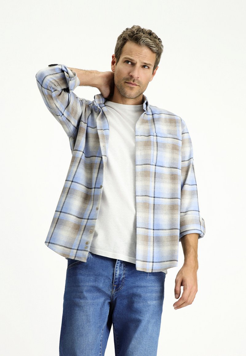 Blauw en bruin geruit flanellen overhemd met knoopsluiting aan de voorkant, opgerolde mouwen, gedragen over een wit T-shirt, gecombineerd met blauwe spijkerbroek. Model poseert casual.