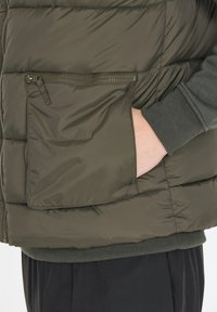 Dunkelgrüne isolierte Weste mit einer Taschen mit Reißverschluss, Steppmuster und einem gerippten Pulloverbund, der sichtbar darunter hervorschaut. Weiche, glatte Textur.