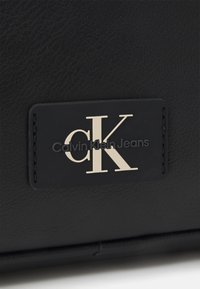 Superficie in pelle nera con un'etichetta rettangolare cucita che mostra le iniziali bianche "cK" e il testo "Calvin Klein Jeans" in nero.