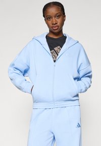 Svetlomodrá zipsová mikina vyrobená z mäkkej tkaniny, s kapucňou, rebrovanými manžetami a vyšitým logom na hrudi. Vhodná s ladícími nohavicami.