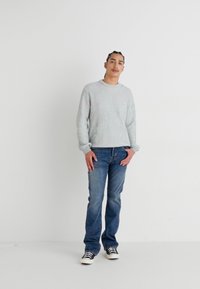 Szürke bordázott pulóver, kerek nyakkivágással, kék farmernadrággal és fekete-fehér sneaker cipőkkel viselve. A modell egy semleges háttérben áll.