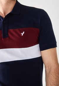 Marineblå, rød og hvit stripete polo skjorte med knappestilling krage; har en brodert fuglelogo på brystet; laget av bomull.