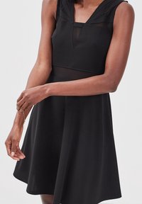 Femme portant une robe noire sans manches avec des panneaux transparents, ajustant un bracelet délicat à son poignet gauche devant un fond uni.