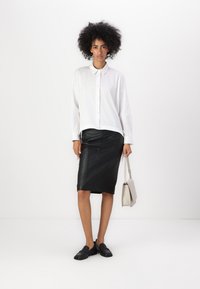 Camicia bianca a maniche lunghe con bottoni, gonna a tubino in pelle nera e mocassini neri. Tiene una borsa grigia chiara, texture morbide, design classico.