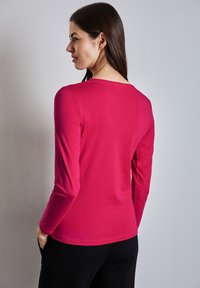 Maglietta a maniche lunghe di un rosa brillante, con scollatura a barchetta e tessuto liscio. Il modello la indossa con pantaloni neri. Design minimalista, senza motivi.