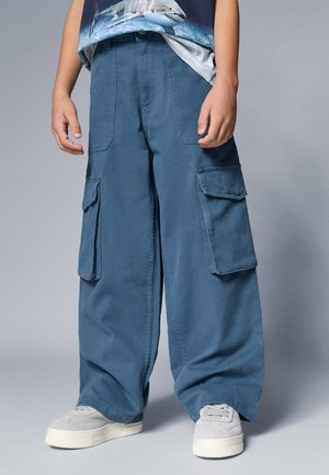 Blaue Cargo-Hosen aus Baumwolle mit großen Seitentaschen, einer lässigen Passform und einer glatten Textur, kombiniert mit hellgrauen Sneakers.