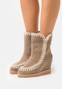 Botas de tobillo con cuña en ante topo con ribete de tela crema. Cuenta con una suela texturizada y costuras decorativas a lo largo del borde.