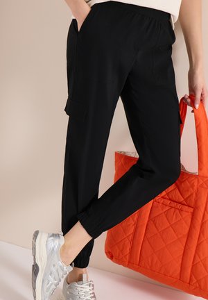Personne portant un pantalon cargo noir et des baskets argentées, tenant un sac fourre-tout matelassé orange sur un fond beige.