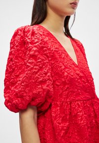 Raudona suknelė su tekstūruotu gėlių raštu, giliu V formos iškirpimu ir pūstomis rankovėmis. Audinys atrodo prabangus ir daugiabriaunis, sukuriantis sluoksniuotą vaizdą.