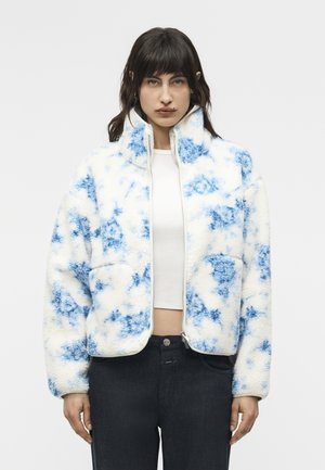 Kobieta w białej i niebieskiej polarowej kurtce w wzory, na białym krótkim topie i czarnych spodniach, stojąca na tle jednolitego szarego tła.