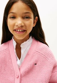 Gilet tricoté rose clair avec un col en V et une fermeture par boutons, porté sur une chemise blanche à col. Présente un petit logo sur la poitrine.