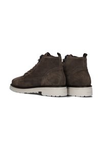 Bruine suède enkelboots met een vetersluiting, witte rubberen zool, versterkte hak en een onderscheidend profiel op de onderzijde.