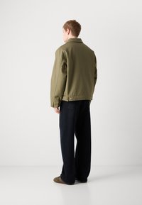 Veste en coton vert olive avec une coupe décontractée, coutures minimales et détails à pression, associée à un pantalon foncé à coupe ample et des chaussures marron.