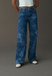 Jeans de mezclilla azules de cintura alta con piernas anchas y un patrón floral. Diseño de cinco bolsillos y cierre de botón metálico, combinados con zapatillas blancas.