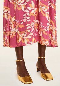 Rochie roz cu imprimeu floral, din material texturat și cu tiv fluid, asociată cu pantofi cu toc bloc aurii și lucioși, prevăzuți cu curele la glezne.