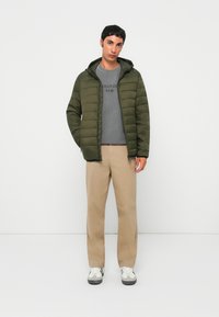 Green padded jacket with a hood, gray sweatshirt featuring text, beige trousers, and white sneakers with black accents, posed against a white background.