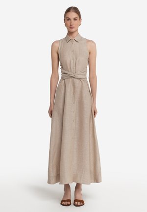 Kvinde iført ærmeløs beige maxikjole med knapper foran, krave og taljebånd, parret med brune åbne sko med hæl, stående mod en hvid baggrund.
