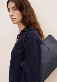 Borsa a tote in pelle blu navy con finitura testurizzata e manici robusti; posizionata accanto a una persona che indossa un maglione scuro, evidenziando il suo design.
