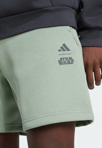 Zelené šortky s logy Adidas a Star Wars, které nosí osoba s tmavou pletí a tmavým tričkem s dlouhým rukávem.