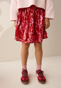 Gonna di paillettes rosse con un motivo scintillante, abbinata a sneakers rosse e calzini rosa. La gonna ha una vita arricciata e una forma svasata.