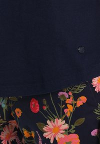 Námořnická modrá halenka s kulatým lemem a černým knoflíkem, spárovaná s živě květinovým vzorem v růžových, oranžových a zelených tónech.