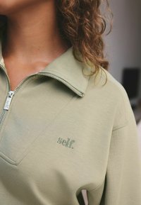 Světle zelená zipová bunda s přeloženým límcem, na které je subtilní vyšívané logo "self." a hladká textilie.