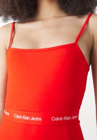 Czerwona bawełniana bluzka na ramiączkach z wąskimi paskami i dopasowanym krojem. Posiada kontrastującą białą taśmę z logo "Calvin Klein Jeans" w pasie.