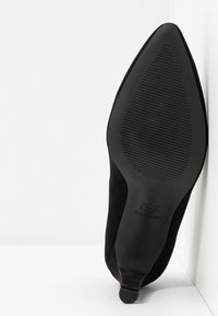 Chaussure à talon haut noire à bout pointu, inclinée vers le haut, montrant la semelle texturée et un petit talon sur fond blanc.