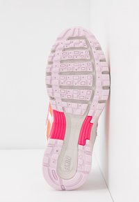 Semelle de basket Nike rose et blanche avec motif de bande de roulement texturé et accents rouges, positionnée verticalement contre un mur d'angle blanc.