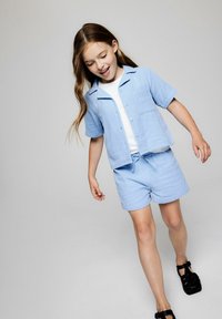 Lyseblå kortærmet skjorte med brystlomme, parret med matchende shorts. Glat tekstur med en afslappet pasform, båret med sorte sandaler.