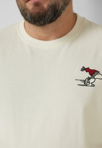 Bärtiger Mann trägt ein cremefarbenes Sweatshirt mit einer kleinen bestickten Snoopy-Figur mit roter Mütze auf der Brust und fährt Schlittschuh.