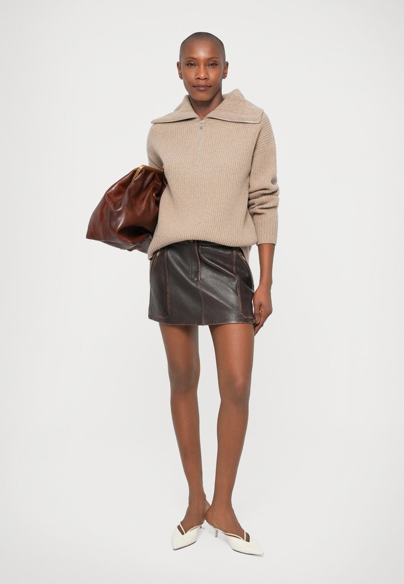 Beige ribbad stickad tröja med dragkedja i kragen, i kombination med en svart läder mini kjol och vita spetsiga klackskor, hållande en brun väska.