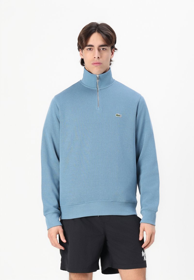 Sweatshirt bleu deschis cu fermoar la gât, textură cu striații, mâneci lungi și tiv cu manșete. Prezintă un mic logo verde pe piept.