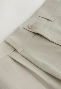 Bež prilagojene hlače s teksturiranim tkanjem, vidnim zapiranjem na gumbe in ravnim sprednjim delom z nežnimi gubami.