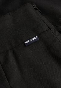 Schwarzer Stoff mit einer glatten Textur, auf einer Tasche ist ein kleines blau-weißes "Superdry"-Etikett aufgenäht. Das Design ist minimalistisch und funktional.