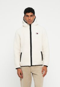 Pehmeä valkoinen fleece-takki mustalla vetoketjulla ja reunoilla, hupulla ja sivutaskuilla. Takissa on pieni, värikäs logo rinnassa.