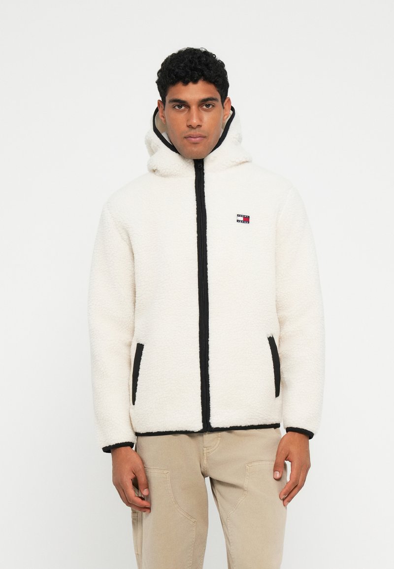 Pehmeä valkoinen fleece-takki mustalla vetoketjulla ja reunoilla, hupulla ja sivutaskuilla. Takissa on pieni, värikäs logo rinnassa.