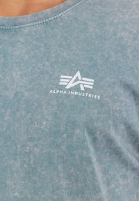 Heled sinise tooni retrostiilis kangas, millel on valge Alpha Industries logo ja tekst rinnapiirkonnas ümarat kaelust.