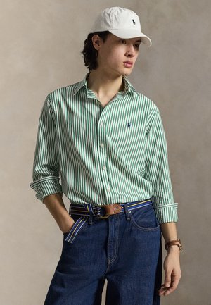 Jauns vīrietis, valkājot baltu cepuri, zaļu un baltu svītrainu pogas krekliņu, zilas džinsas un zilu svītrainu jostu, stāv ar vienu roku kabatā.