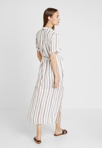 Vestido maxi com colarinho, mangas curtas e cintura com cinto. Apresenta riscas verticais brancas e castanhas, fendas laterais e tecido leve.