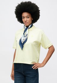 Γυναίκα με αφρο μαλλί, φορώντας ανοιχτό κίτρινο πόλο, ναυτικό φλοράλ μαντήλι δεμένο γύρω από τον λαιμό και σκούρο μπλε τζιν, στέκεται με το ένα χέρι στην μέση.