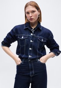 Femme aux cheveux roux courts portant une veste en jean bleu foncé assortie à un jean taille haute, les mains dans les poches, sur un fond clair uni.